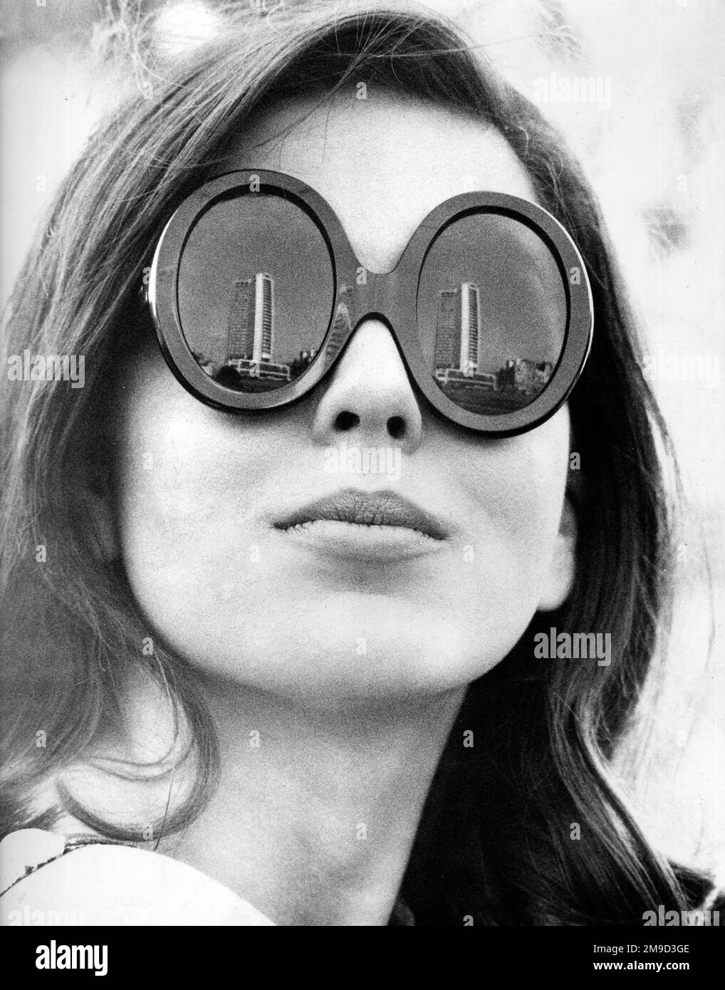 Weibliches Model trägt eine Sonnenbrille mit dickem Rahmen, wobei sich ein Wolkenkratzer-Gebäude in den Gläsern spiegelt. Stockfoto