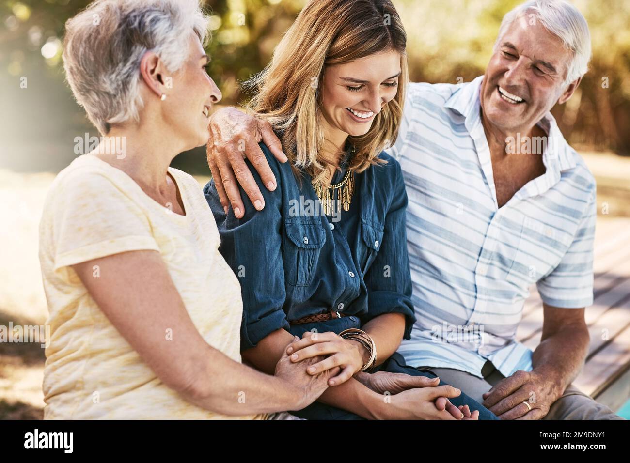 Ältere Paare in der Natur mit ihrer erwachsenen Tochter, die in einem Garten sitzt, sich unterhält und sich zusammenhält. Glückliche, liebe und ältere Menschen, die das Kind umarmen Stockfoto