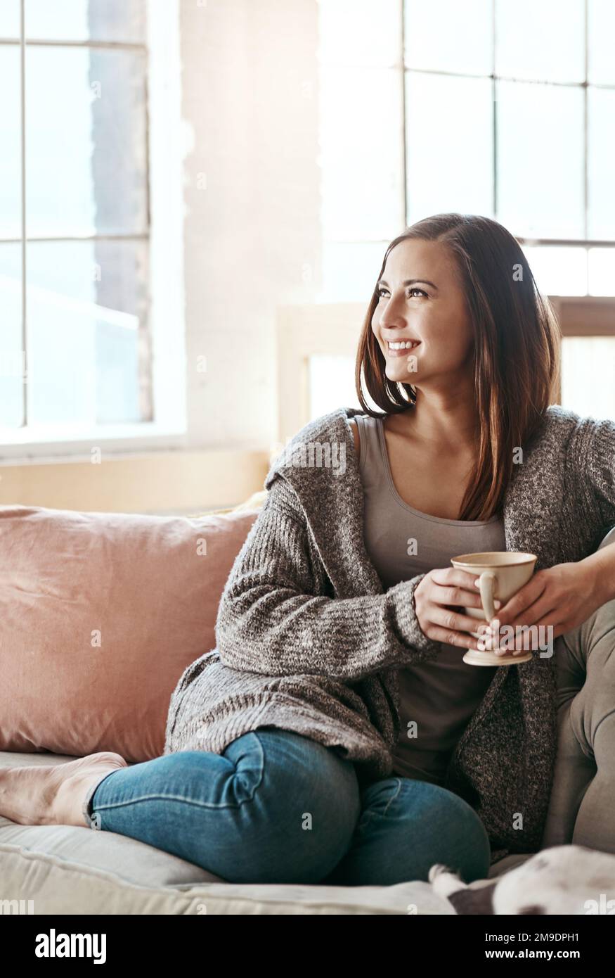 Entspannen Sie sich, Kaffee und Frau auf einer Wohnzimmercouch und fühlen Sie sich wie zu Hause. Tee, Glück und Lächeln einer Person am Morgen, mit der man nachdenkt Stockfoto