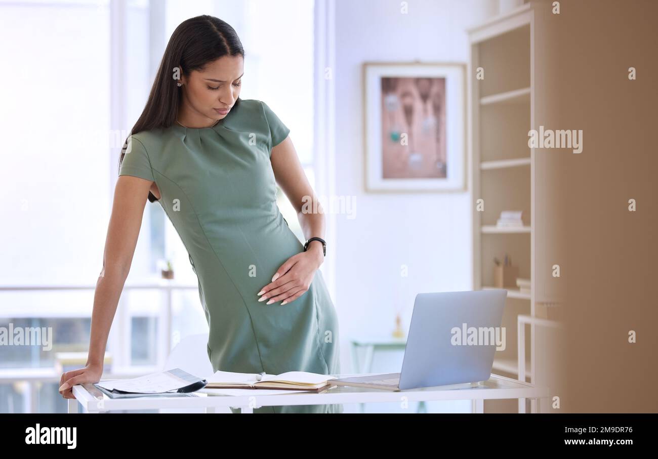 Ich glaube, ich muss zu meinem Arzt. Eine junge schwangere Geschäftsfrau, die allein im Büro steht und unter Bauchschmerzen leidet. Stockfoto
