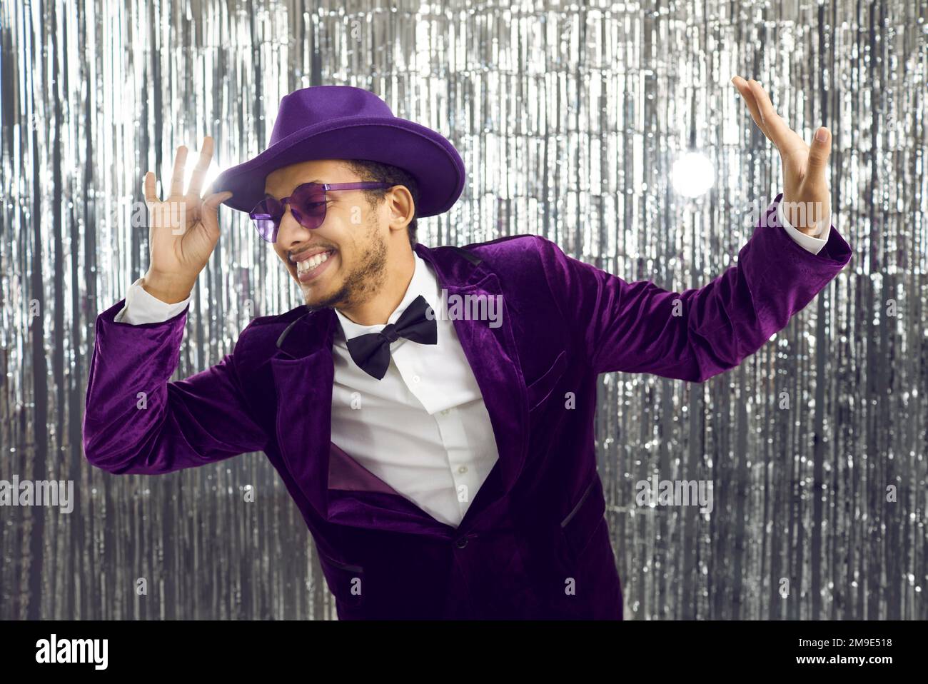 Fröhlicher, lustiger junger Kerl in einem lila Anzug, Brille und Hut, tanzend und Spaß auf einer Party zu haben Stockfoto