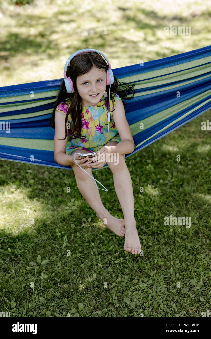 Mädchen, 8 Jahre, sitzt in einer Hängematte im Schatten und hört Musik mit Kopfhörern Stockfoto