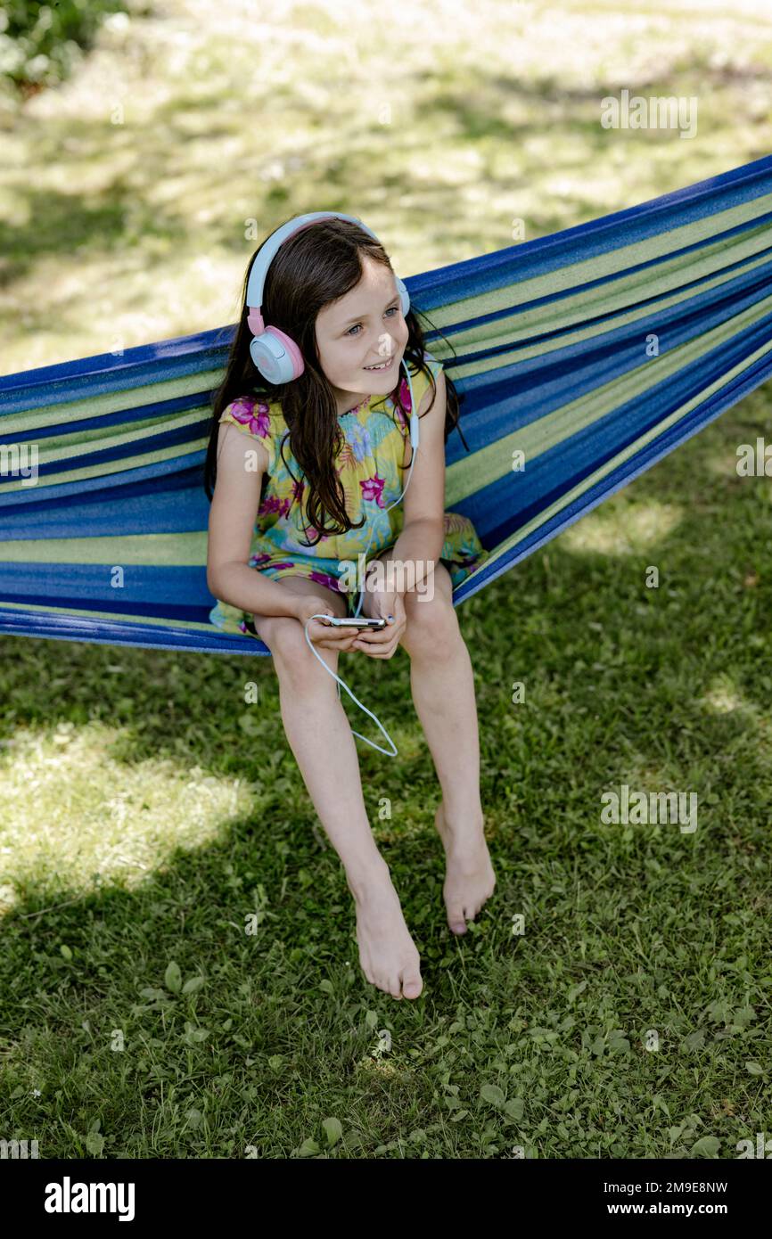 Mädchen, 8 Jahre, sitzt in einer Hängematte im Schatten und hört Musik mit Kopfhörern Stockfoto