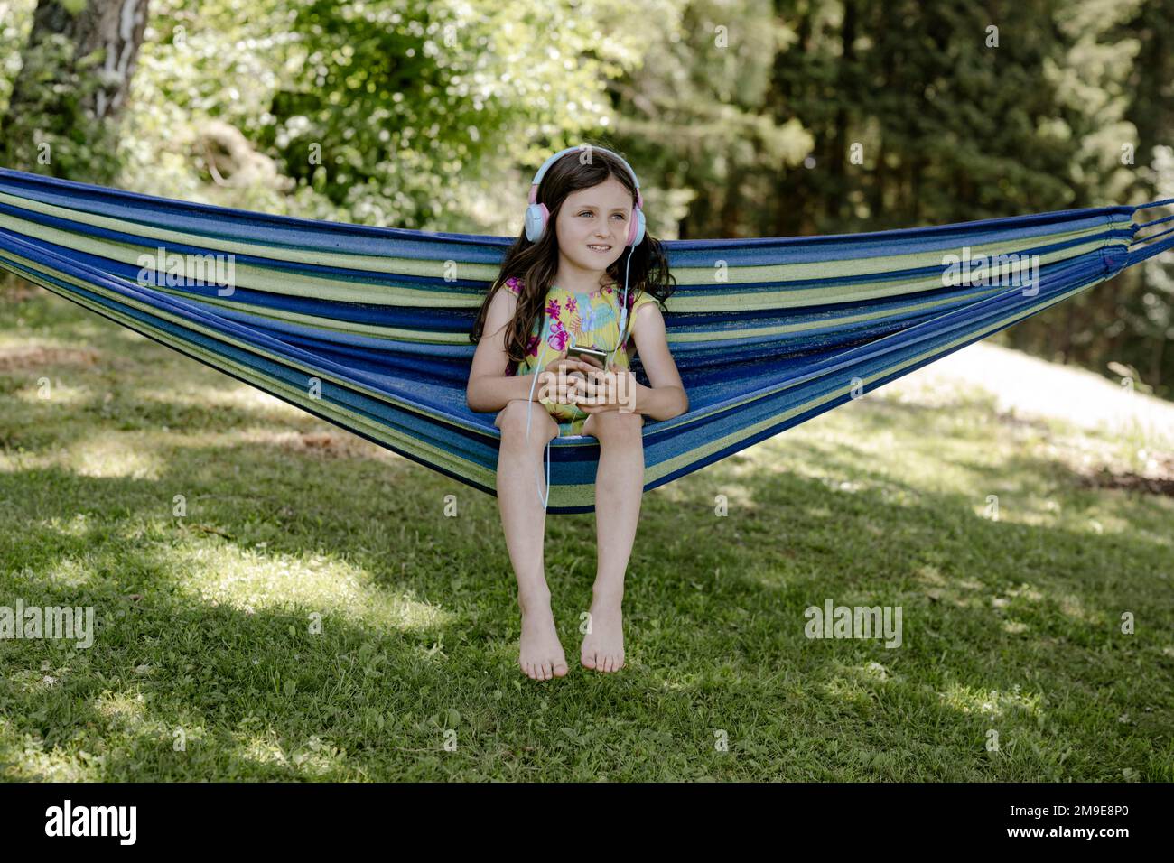 Mädchen, 8 Jahre, sitzt in einer Hängematte im Schatten und hört Musik mit Kopfhörern Stockfoto