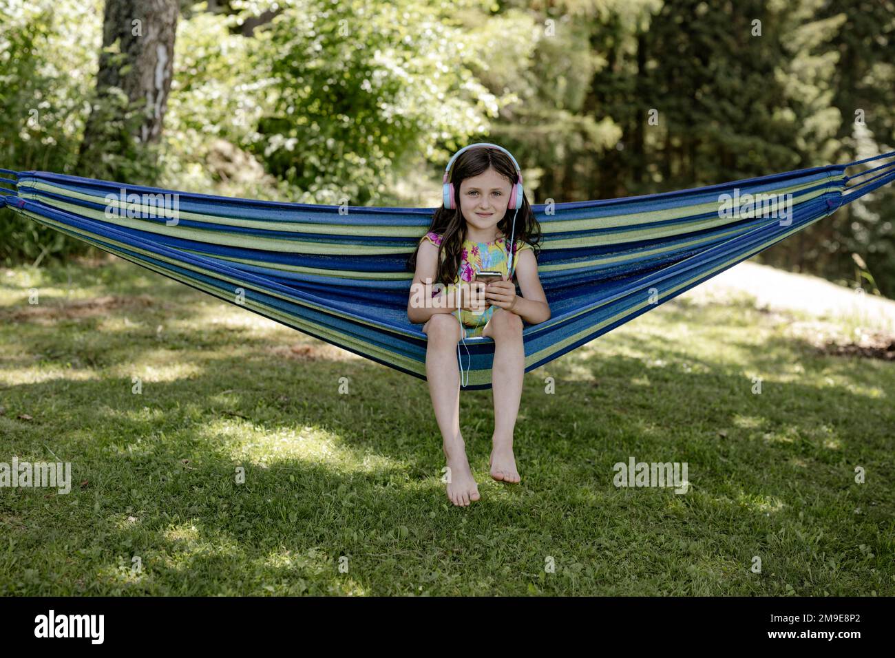 Mädchen, 8 Jahre, sitzt in einer Hängematte im Schatten und hört Musik mit Kopfhörern Stockfoto