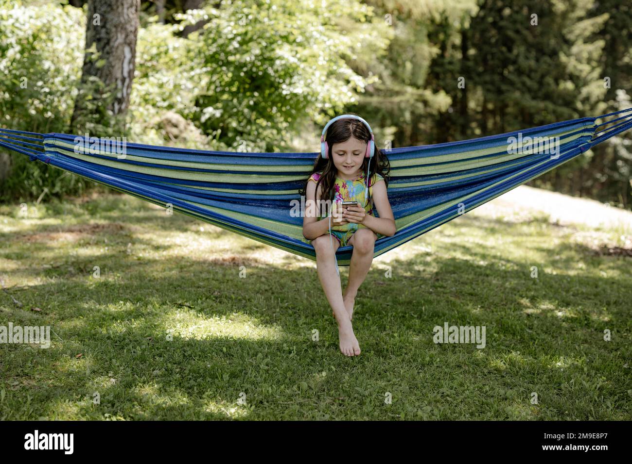 Mädchen, 8 Jahre, sitzt in einer Hängematte im Schatten und hört Musik mit Kopfhörern Stockfoto