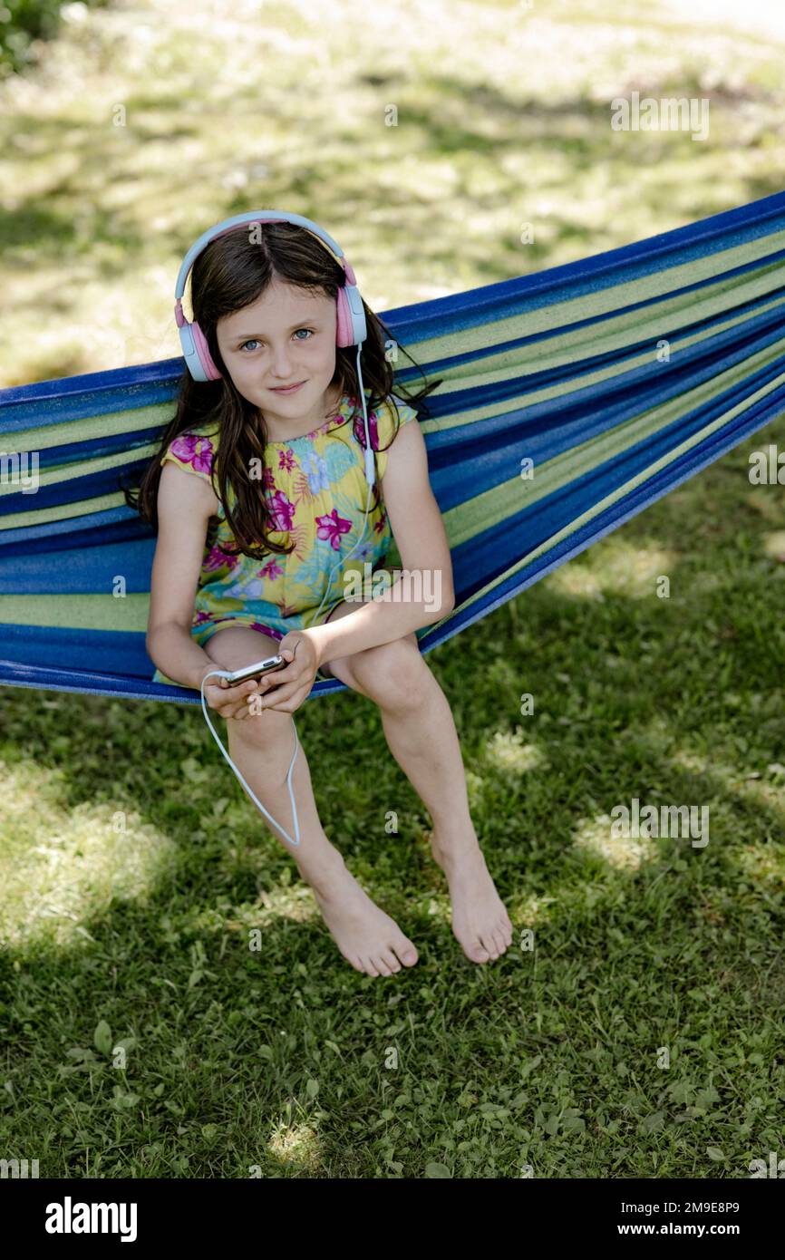 Mädchen, 8 Jahre, sitzt in einer Hängematte im Schatten und hört Musik mit Kopfhörern Stockfoto
