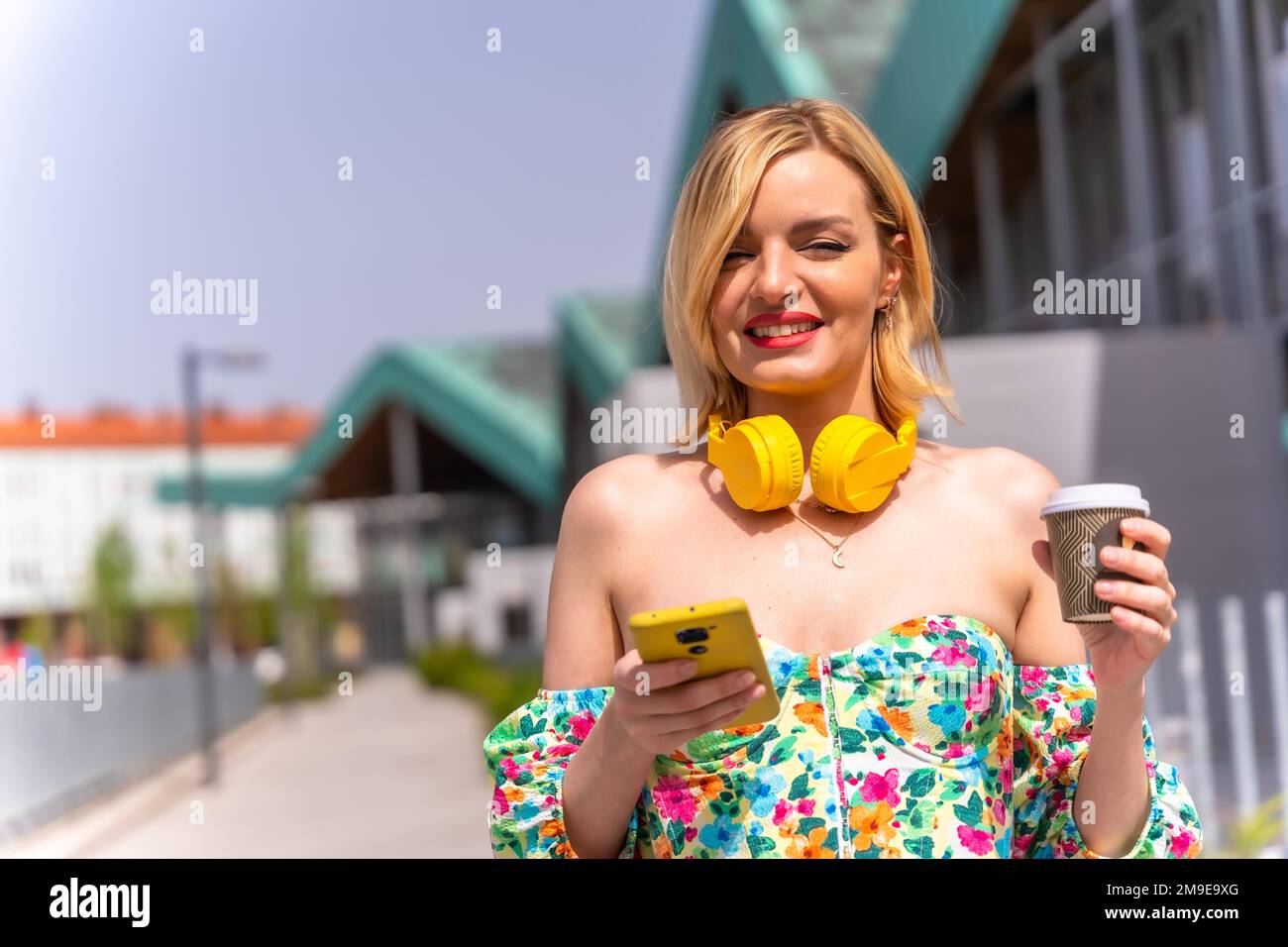 Porträt einer blonden Frau, die mit dem Telefon durch die Stadt läuft, mit gelben Kopfhörern und einem Kaffee zum Mitnehmen, Lebensstil eines Models in der Stadt Stockfoto