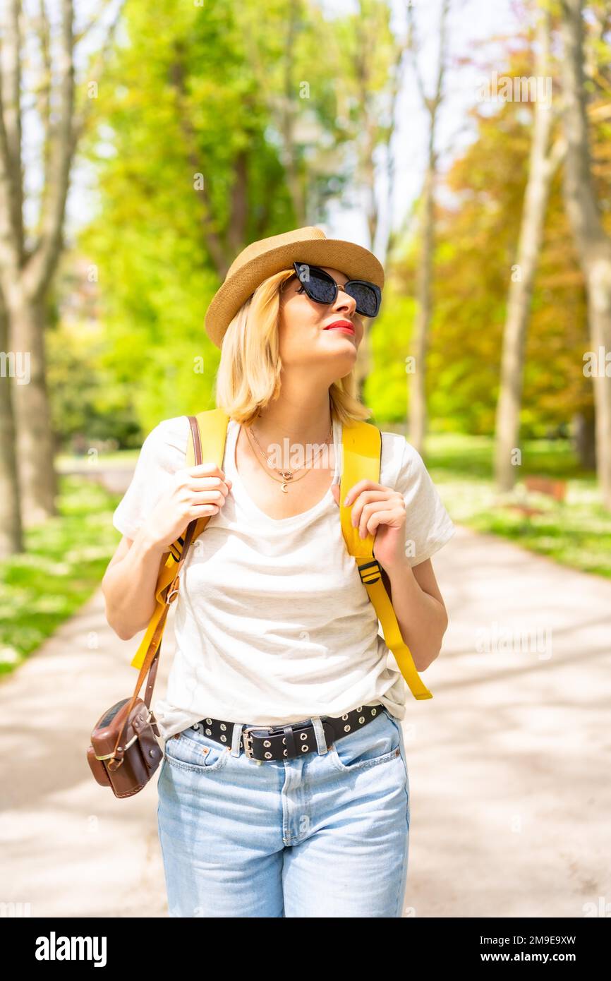 Eine touristische blonde Frau mit Hut und Sonnenbrille, die im Frühling in einem Park in der Stadt mit der Kamera spaziert und die Sonne genießt Stockfoto