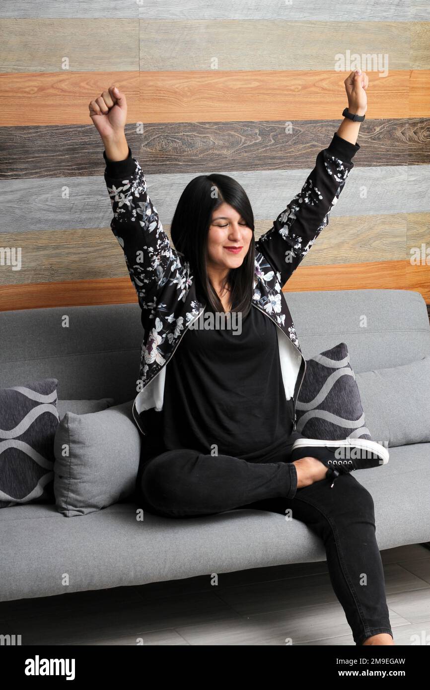 Lateinische Erwachsene Frau, die auf dem Sofa sitzt, ist ein Zeichen für Sieg, Freiheit und Glück für den finanziellen Erfolg ihres Unternehmens und ihres vollen Lebens Stockfoto
