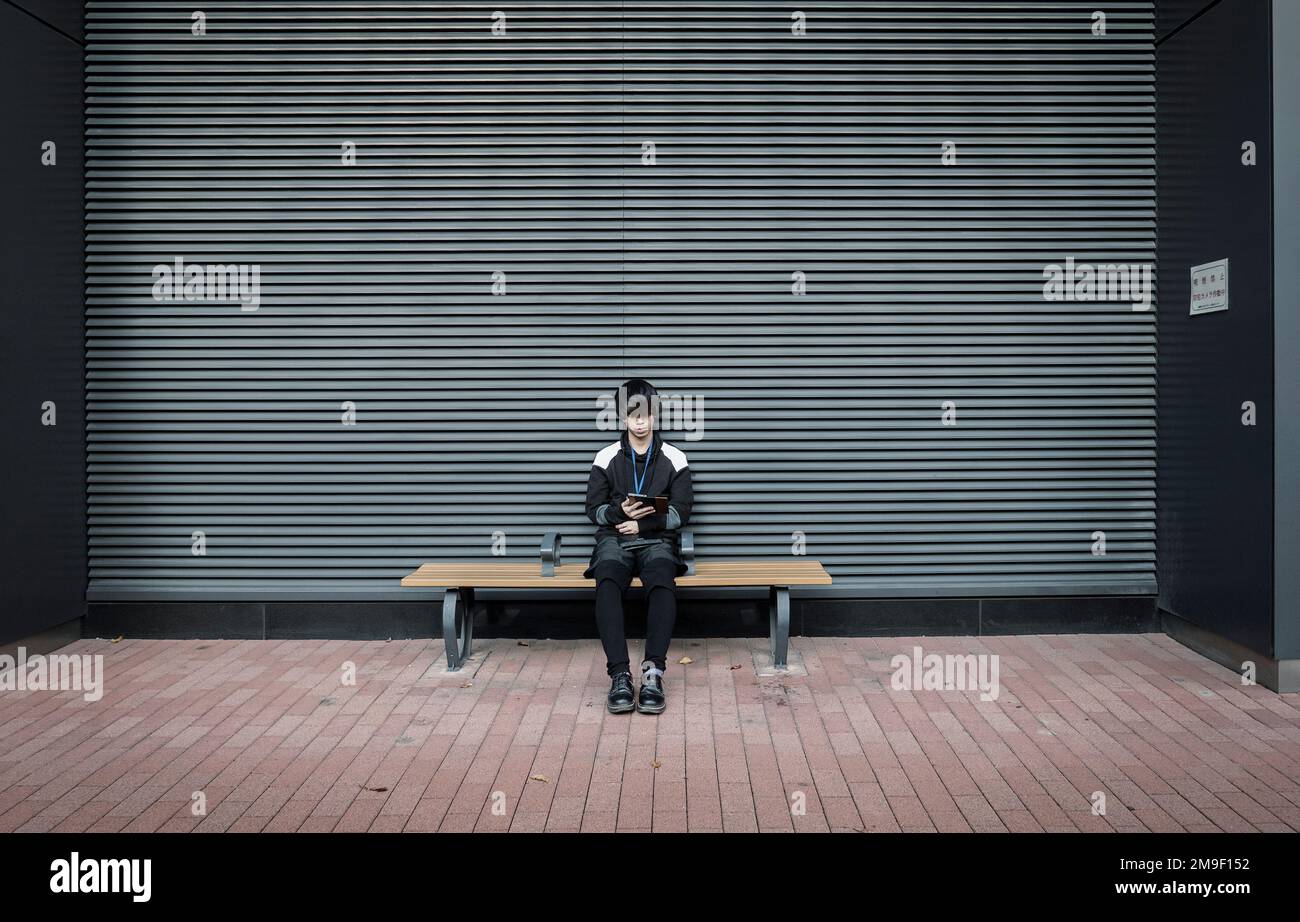 Tokio - Japan - Ca. November 2018. Ein einsamer Junge, der mit dem Smartphone gegen einen geschlossenen Verschluss interagiert. Einsamkeitskonzept. Stockfoto