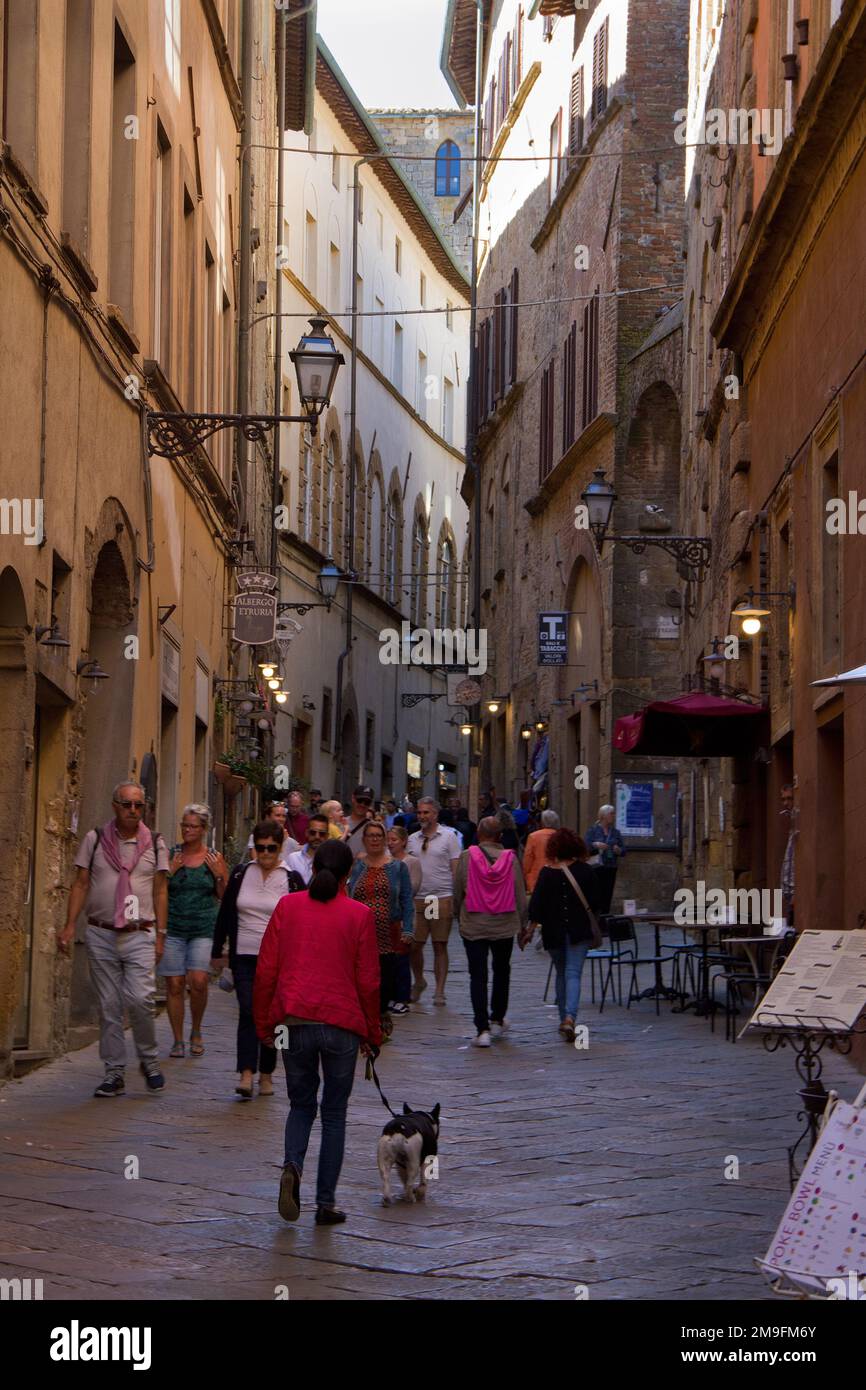 Touristen (nicht wiederzuerkennen), die eine schattige, enge, lebhafte Straße in einer Stadt in der Toskana entlangspazieren Stockfoto