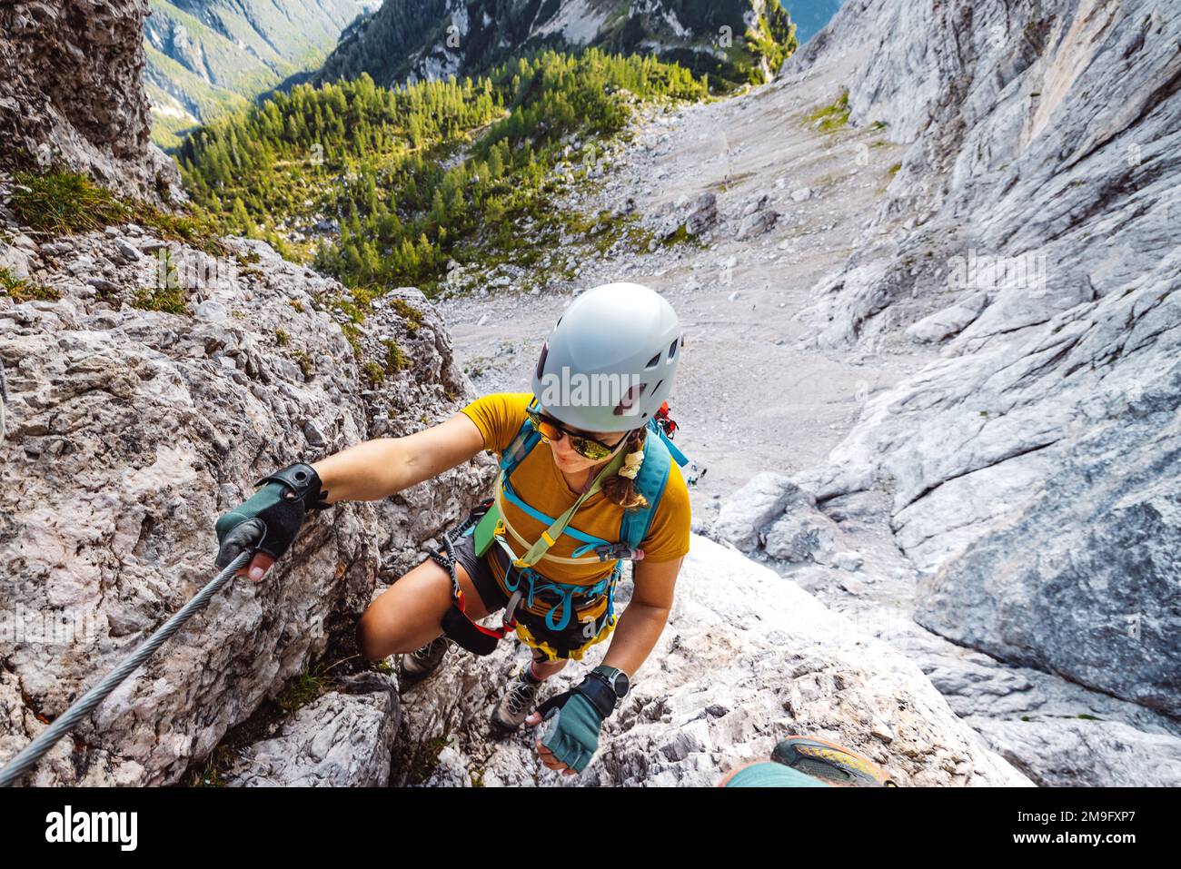 Ich schaue hinunter auf eine weißhäutige Kletterfrau, die hoch oben in den Bergen mit dem Ferrata-Zug hochklettert Stockfoto