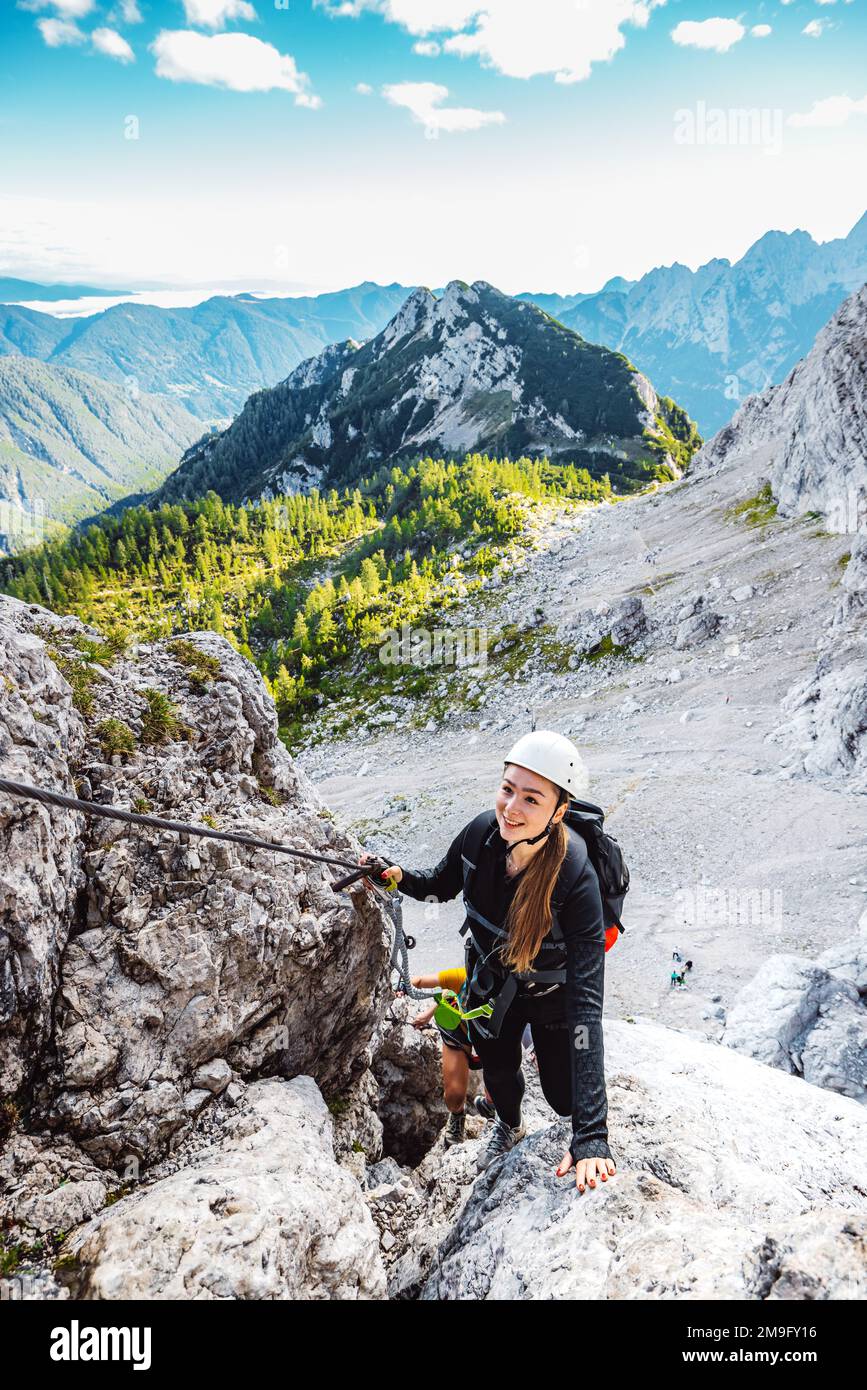 Vertikales Foto einer weißhäutigen Klettererin auf dem Bergsteigerpfad in den Alpen, die mit Helm und Sicherheitsgurt klettert Stockfoto