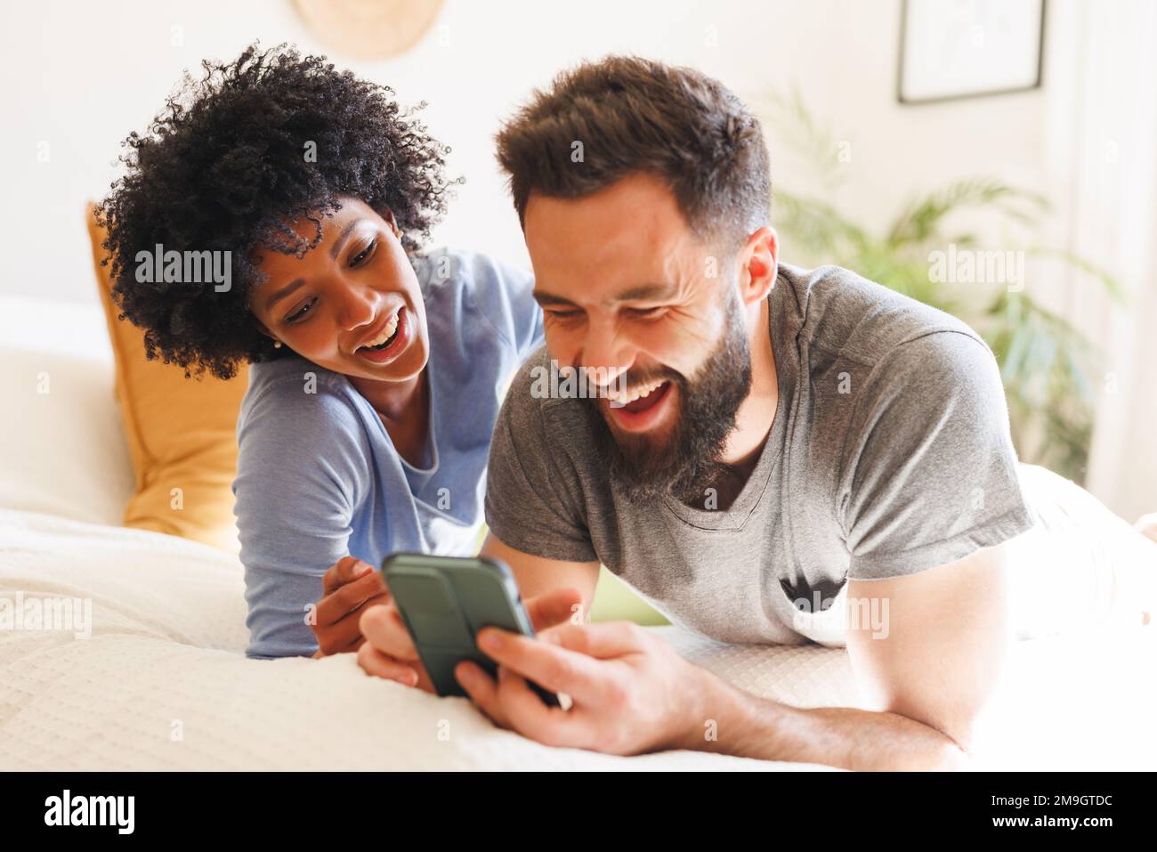 Fröhliches, birassisches junges Paar, das sich ein Smartphone anschaut und lacht, während es im Bett liegt Stockfoto