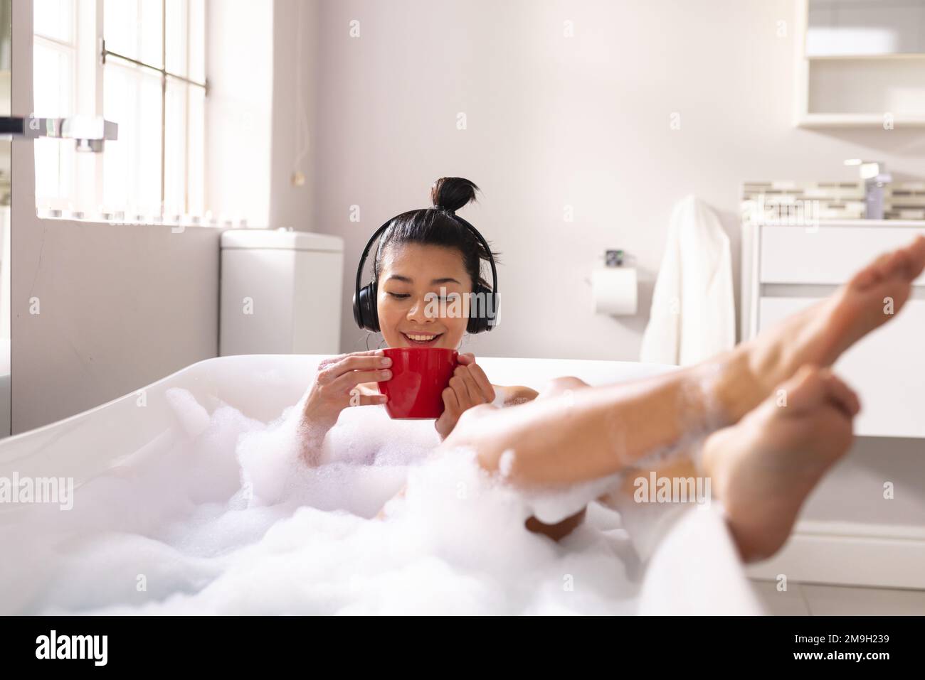 Bild einer birassischen Frau, die sich mit einer Tasse Tee und Kopfhörern in der Badewanne im Schaumbad entspannt Stockfoto