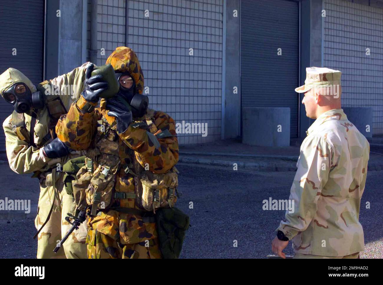 Ein Australier erhält erfolgreich einen Schluck Wasser aus seiner Kantine, während er sich in einer Mission-Oriented Protective Hure Response Level 4 (MOPP-4) Ausrüstung befindet. Der Australier trägt seinen MKIV No1 Nuclear Biological Chemical (NBC) Anzug mit Avon FM12 Maske und His Colt 5,56 mm M4 Carbine. Hinter ihm steht ein Marine in seinem Joint Service Lightweight Integrated Suit Technology (JSLIST), der eine chemisch-biologische Feldmaske M40 mit einem C2A1-Filterkanister trägt, und nimmt Anpassungen an der Ausrüstung der Australier vor. Einschätzer, Sergeant Robert T. Mawson beobachtet den Prozess. Die Marines und Australier nehmen an NBC Traini Teil Stockfoto