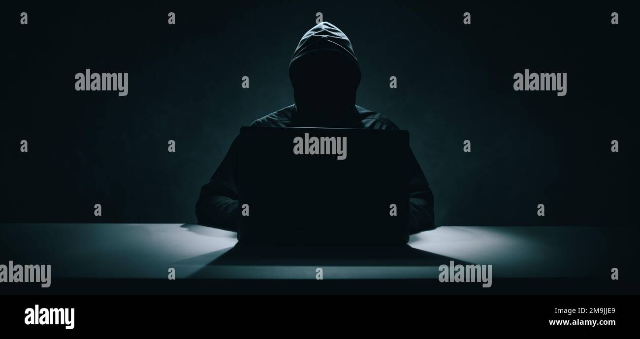 Hacker in schwarzem Hoodie, der im Dunkeln an einem Laptop arbeitet. Cybersicherheit. Banner Stockfoto