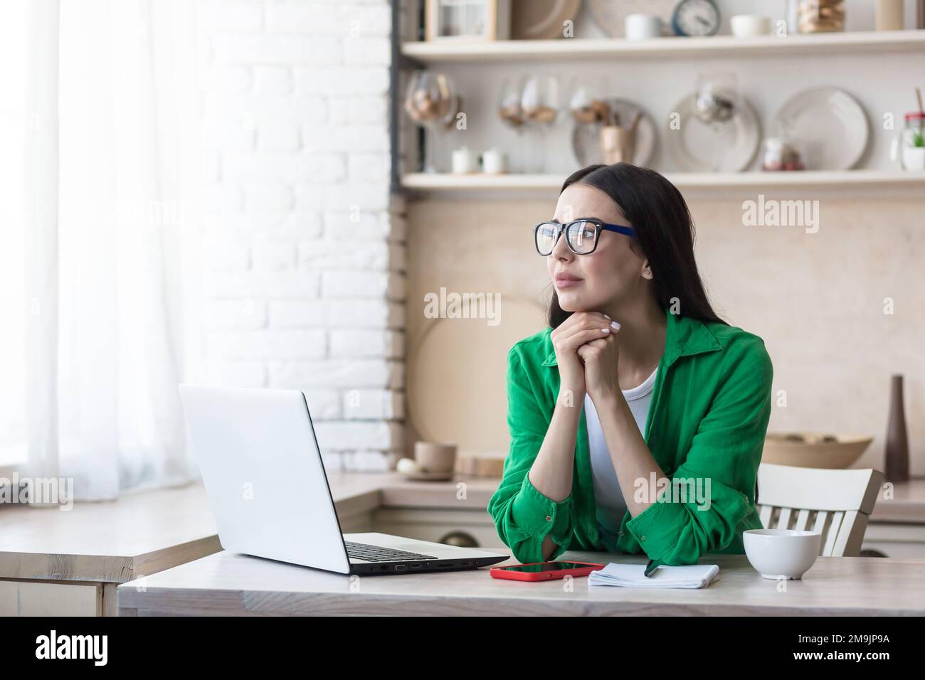 Verärgerte junge Frau, die zu Hause in der Küche vor einem Laptop sitzt. Geben Sie auf der Tastatur ein, suchen Sie, überprüfen Sie. Sie schaut durchdacht aus dem Fenster und legt ihren Kopf auf ihre Hände. Stockfoto