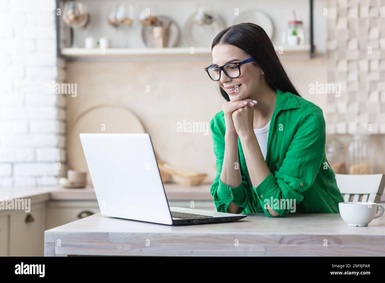 Junge, wunderschöne Bloggerin sitzt zu Hause in der Küche und chattet online mit einer Laptop-Kamera. Sie ruhte ihren Kopf auf ihren Händen aus, hörte zu, erzählte Geschichten, lächelte. Stockfoto