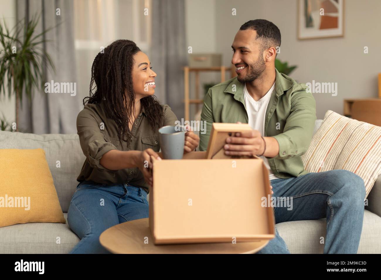 Glücklicher schwarzer Familienvater, Ehemann und Ehefrau, die Sachen aus dem Karton auspacken, nachdem sie umgezogen sind, sitzen auf dem Sofa Stockfoto