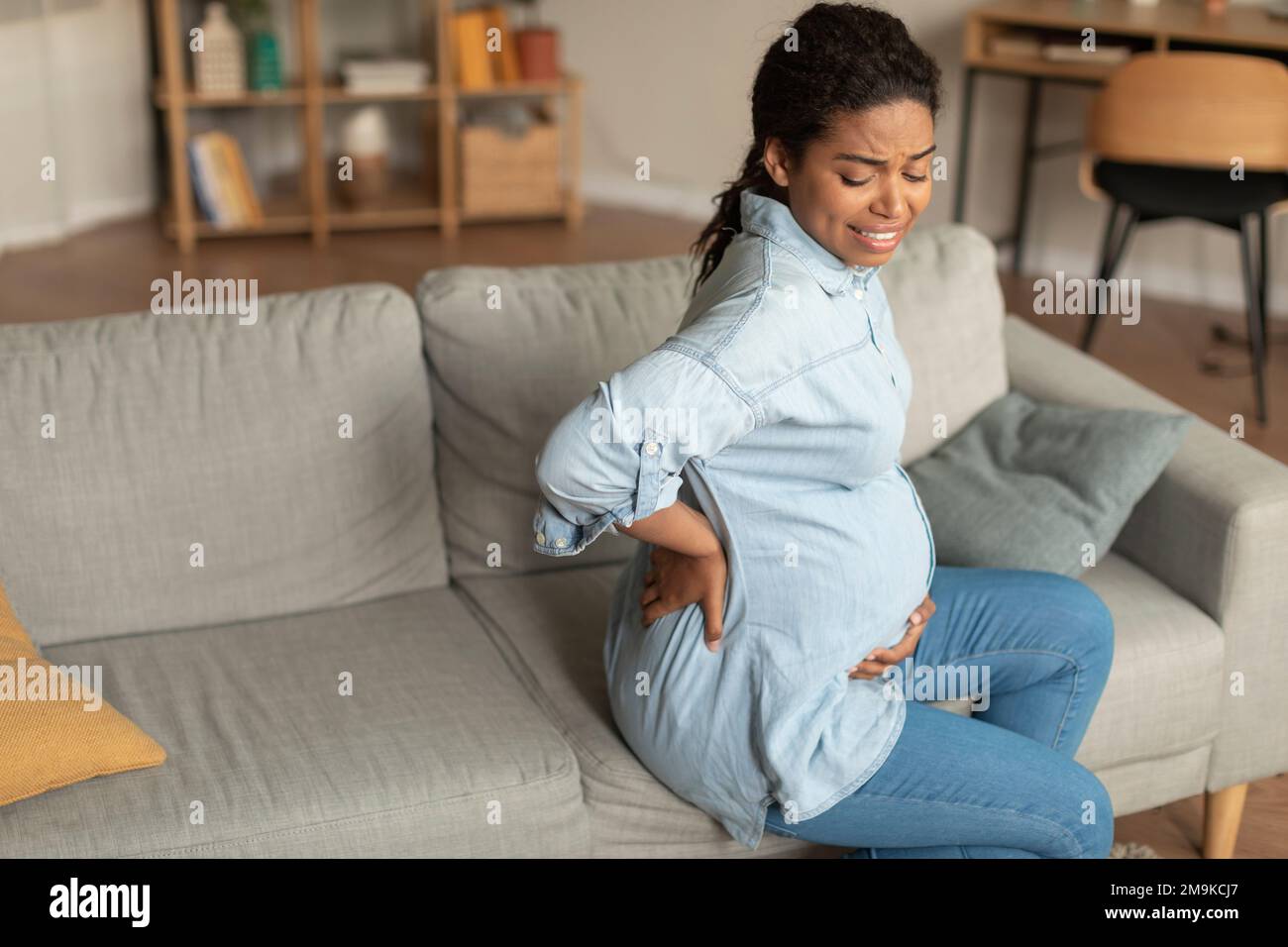 Traurige, schwarze, schwangere Frau mit großem Bauch, die Rückenschmerzen hatte, saß auf dem Sofa im Wohnzimmer, freier Platz Stockfoto