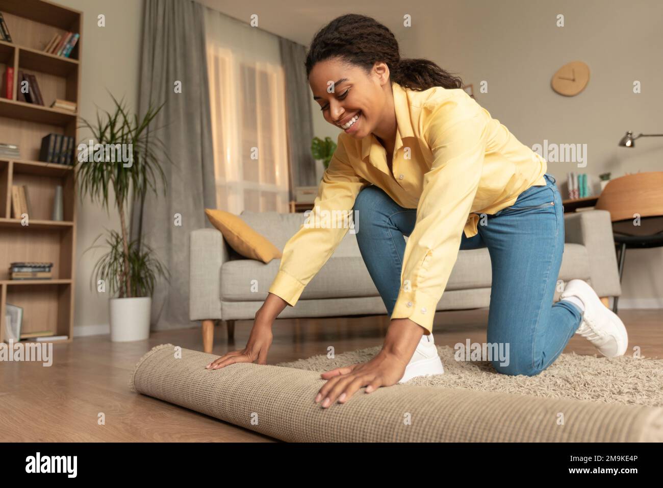 Junge afroamerikanische Frau, die den Haushalt macht, sich bückt und Teppich in einem Wohnzimmer rollt Stockfoto