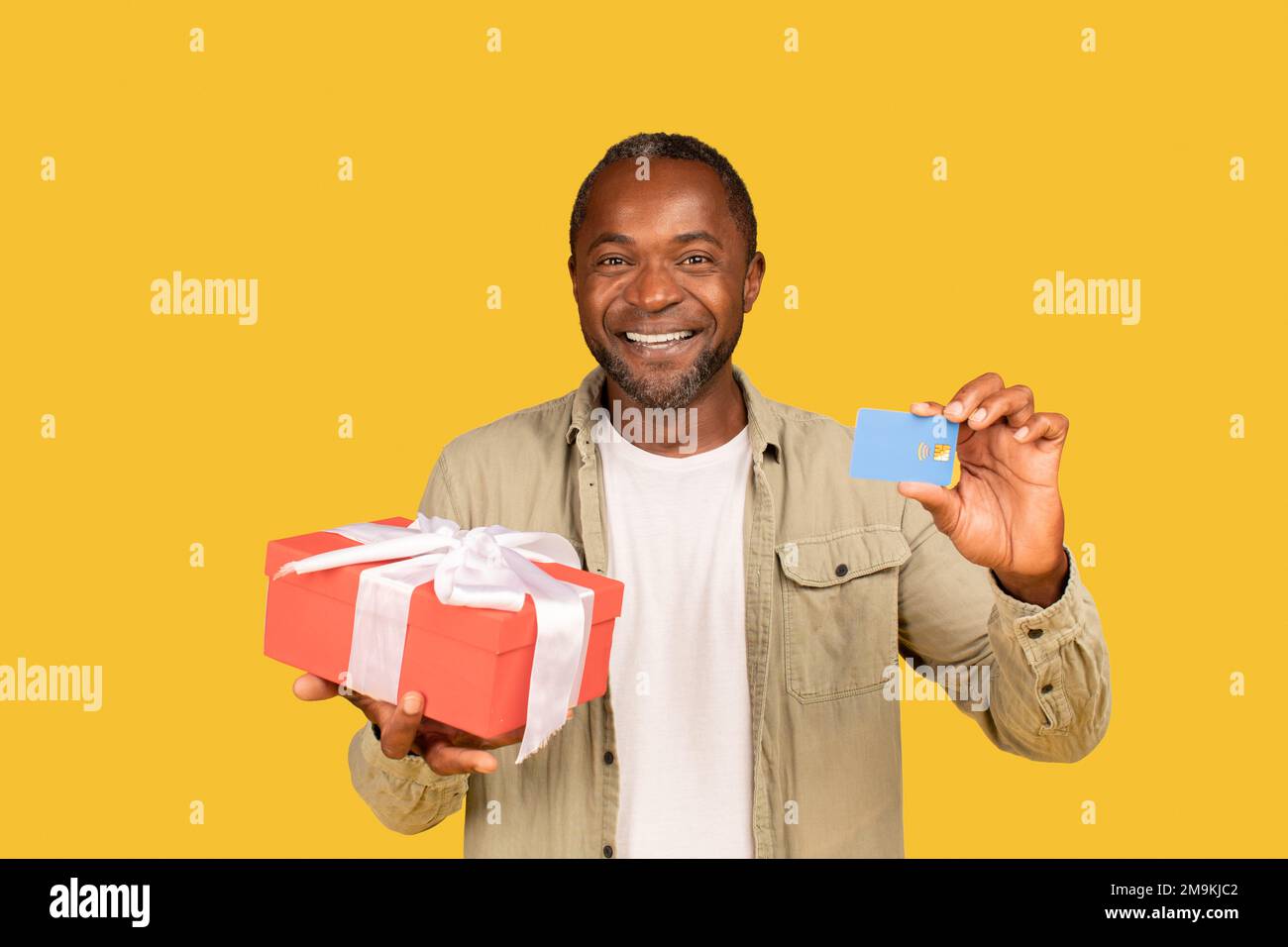 Fröhlicher afroamerikanischer Mann mittleren Alters zeigt Geschenkbox und Kreditkarte, empfehle Bankwesen Stockfoto