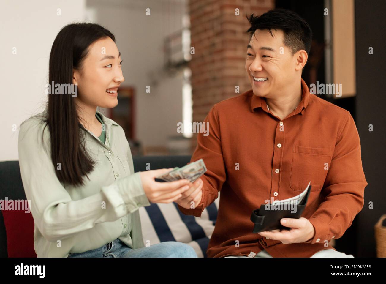 Wohlhabender koreanischer reifer Ehemann, der seiner jungen Frau Geld gibt, sitzt zu Hause auf dem Sofa Stockfoto