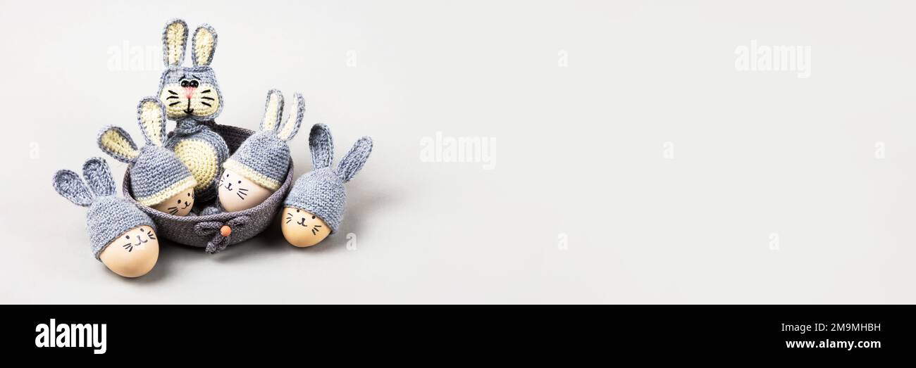 Fünf Ostereier mit lustigen Gesichtern und graue gehäkelte Hüte mit Hasenohren in einem dekorativen Korb und ein gehäkeltes graues Häschen auf grauem Hintergrund. Happ Stockfoto