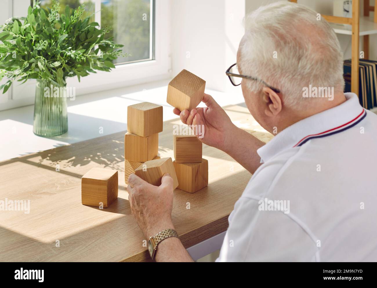Ein älterer Mann sammelt Holzwürfel am Tisch gegenüber dem Fenster. Stockfoto