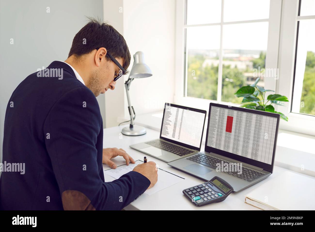 Buchhalter schreibt etwas, während er mit digitalen Blättern auf Bürocomputern arbeitet Stockfoto