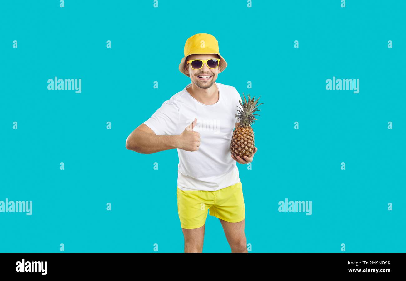 Ein fröhlicher Mann in Sommerkleidung und mit Ananas in der Hand zeigt Daumen nach oben auf hellblauem Hintergrund. Stockfoto