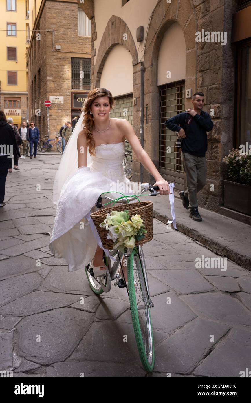 Eine Braut in einem langen Brautkleid fährt mit dem Fahrrad in einer gepflasterten Seitenstraße in Florenz, Italien Stockfoto