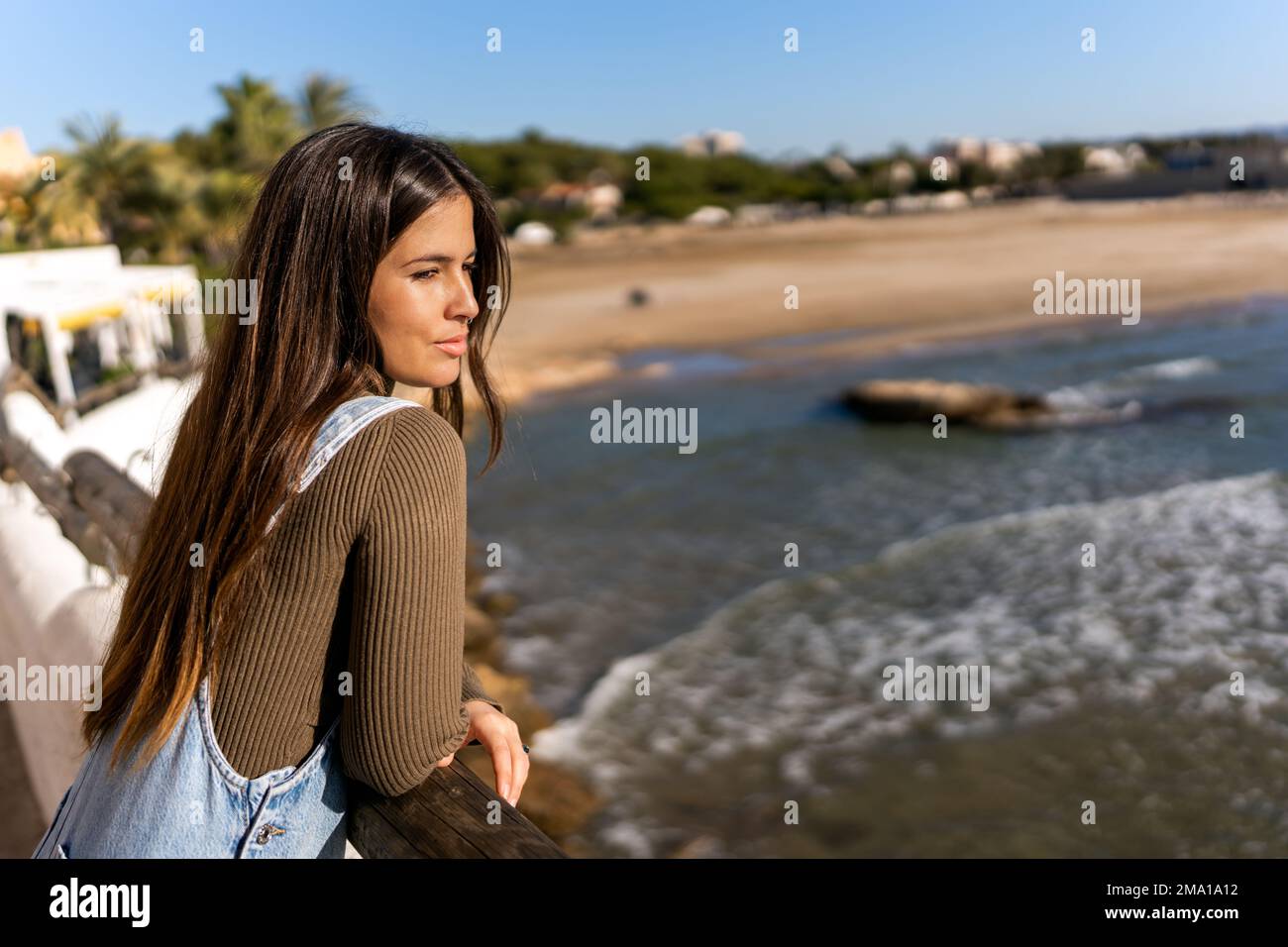Seitenansicht der jungen Frau in legerer Kleidung mit langen dunklen Haaren, die auf dem Geländer liegen, während Sie die malerische Landschaft des Meeres an sonnigen Tagen bewundern Stockfoto