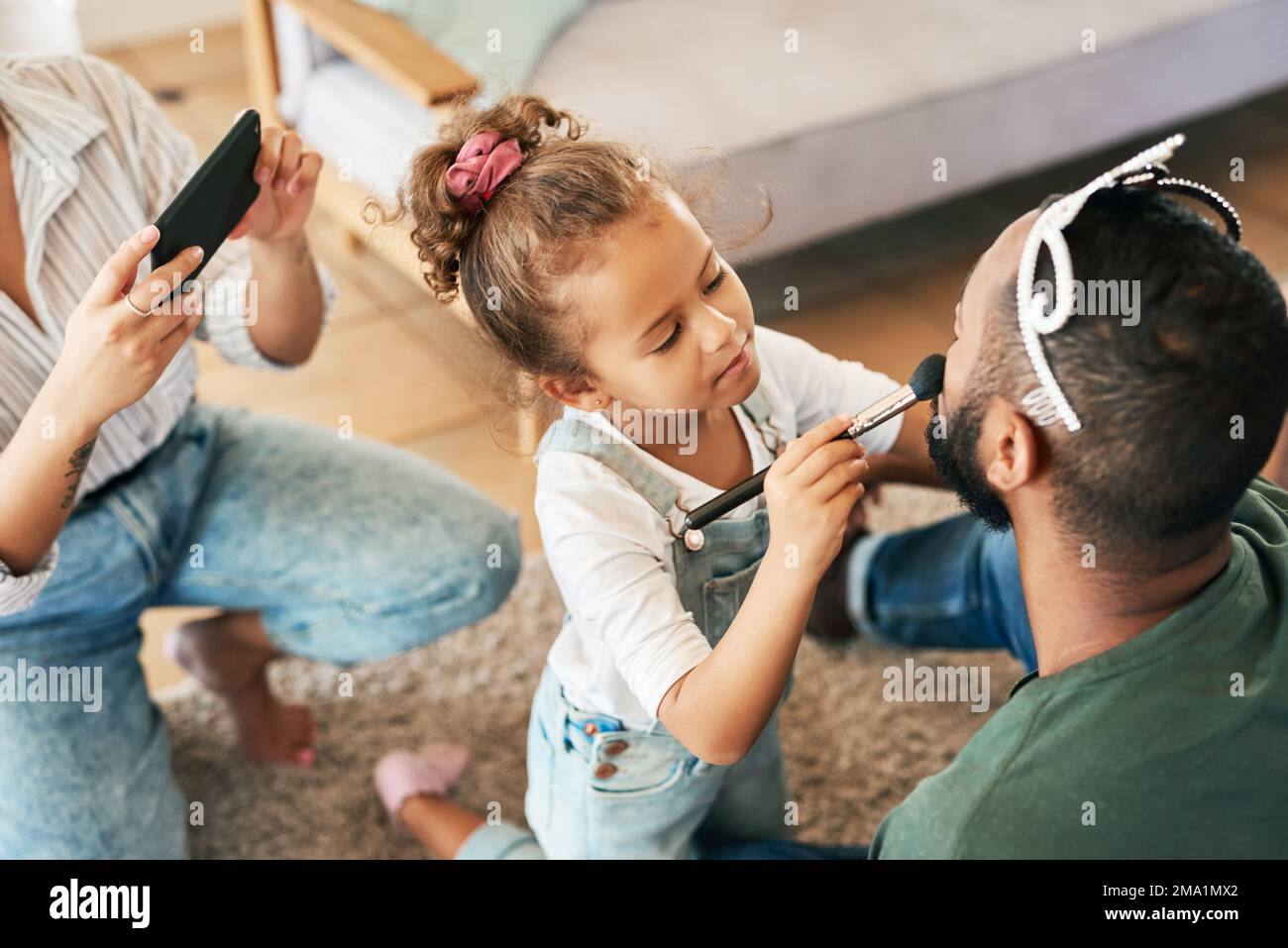 Ein kleiner Beauty-Guru in der Herstellung. Ein kleines Mädchen schminkt ihren Vater, während ihre Mutter zu Hause Fotos von ihnen macht. Stockfoto