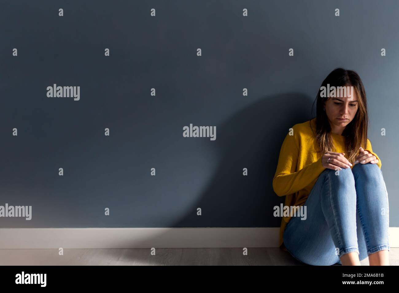 Junge brünette Frau mit Depressionen, die auf dem Boden eines Raumes sitzt und sich an eine graue Wand lehnt Stockfoto
