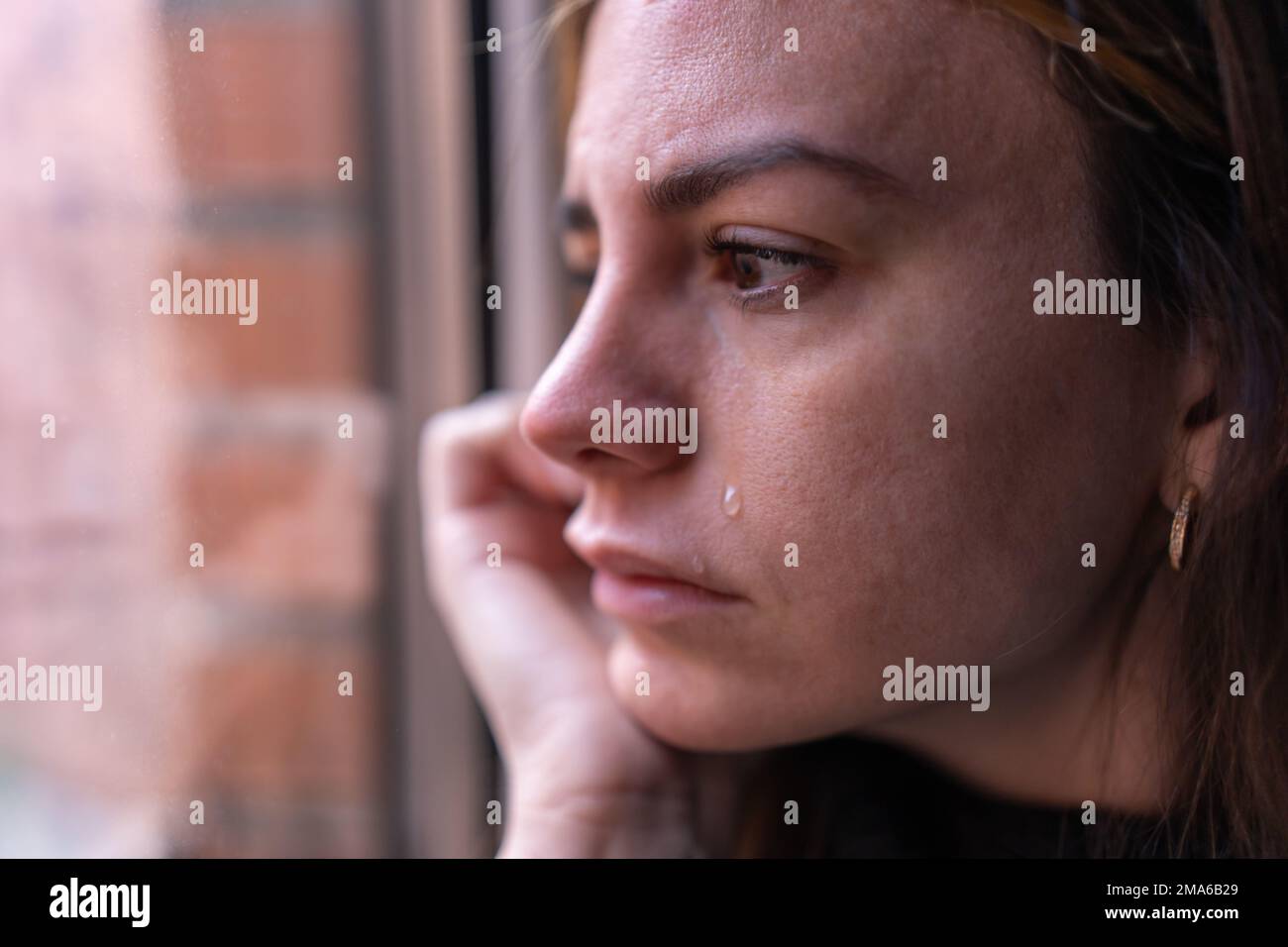 Nahaufnahme einer depressiven Frau, die aus dem Fenster schaut, während eine Träne ihre Wange runterfällt. Depression und psychisches Gesundheitskonzept Stockfoto