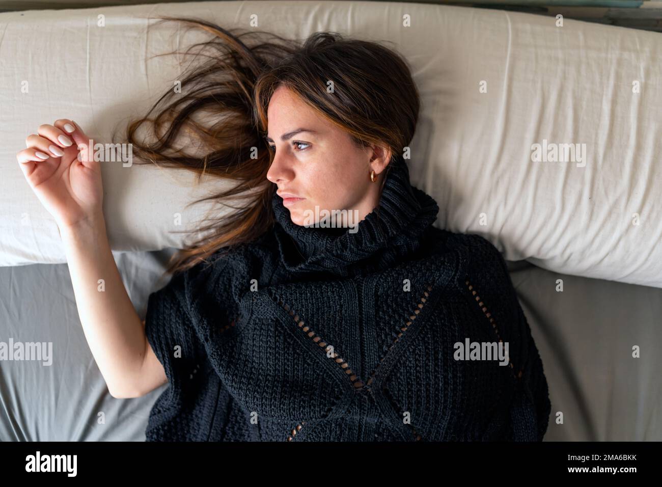 Blick von oben auf eine junge brünette Frau in einem schwarzen Kleid, die traurig aussieht und auf dem Bett liegt. Depression und psychisches Gesundheitskonzept Stockfoto