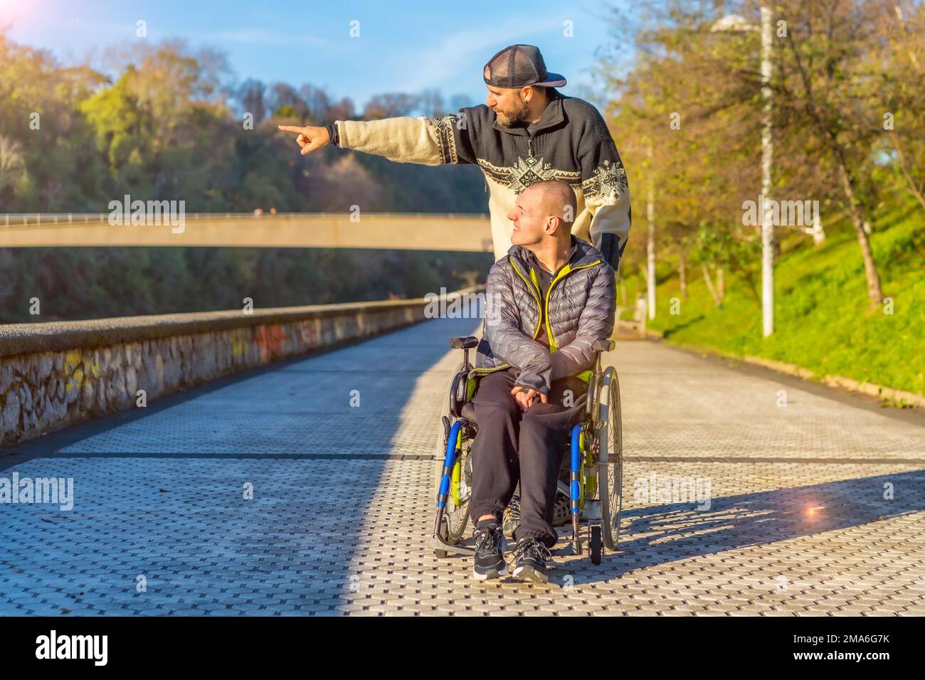 Lächelnde behinderte Person im Rollstuhl, die mit ihrem Bruder bei Sonnenuntergang in einem Park spaziert Stockfoto