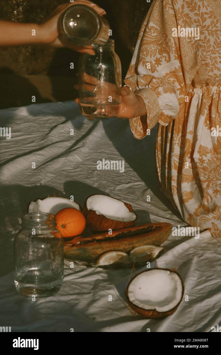 Eine vertikale Aufnahme einer weiblichen Hand, die Wasser in ein Glasgefäß in die Hand einer anderen Frau gießt, und es gibt Kokosnuss mit Orangenfrucht auf einem Laken unter ihren Händen Stockfoto