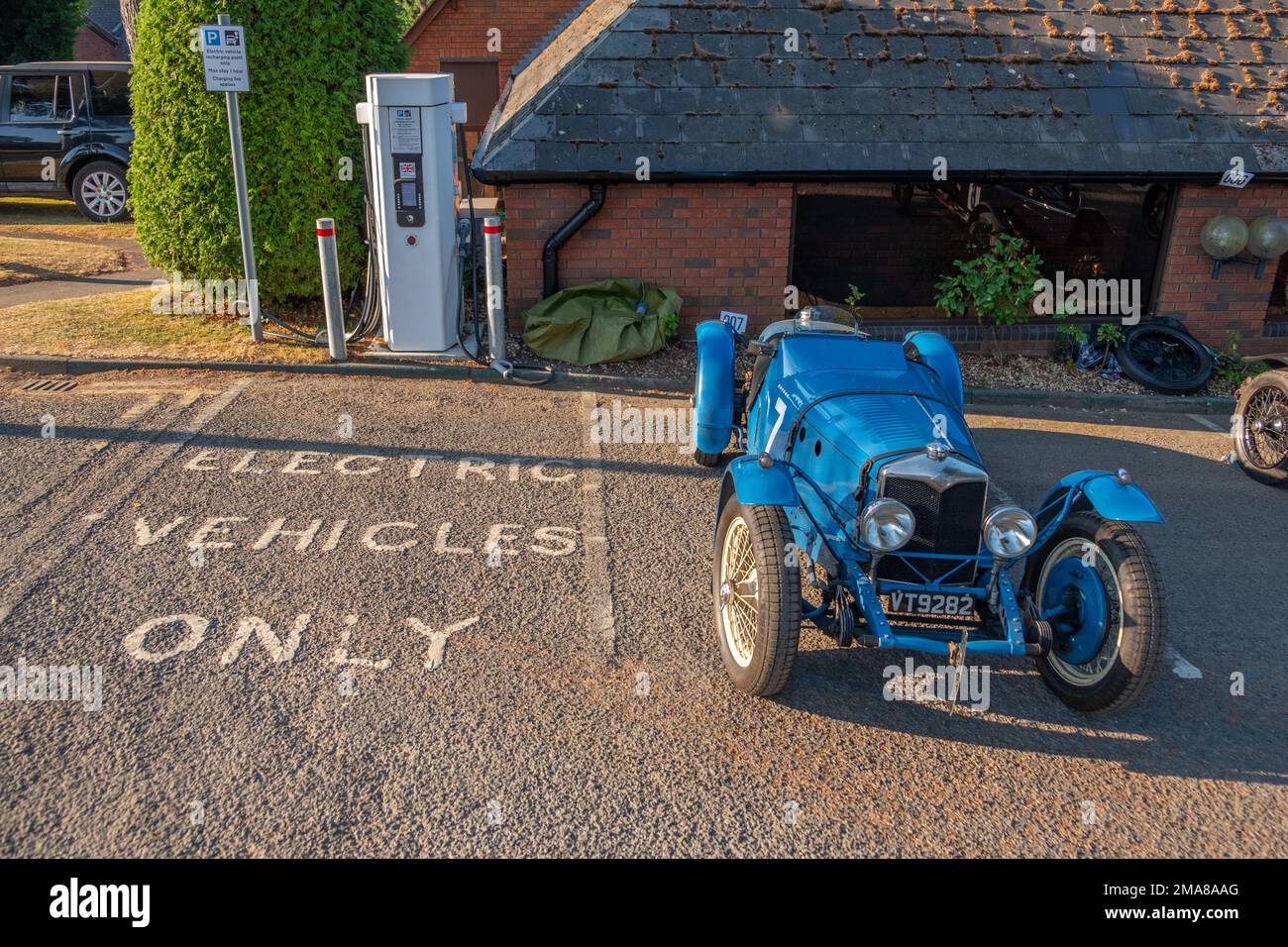 Blaues 1933 Riley TT Sprite Classic Auto neben einem Ladepunkt für Elektrofahrzeuge bei niedriger Morgensonne. Stockfoto