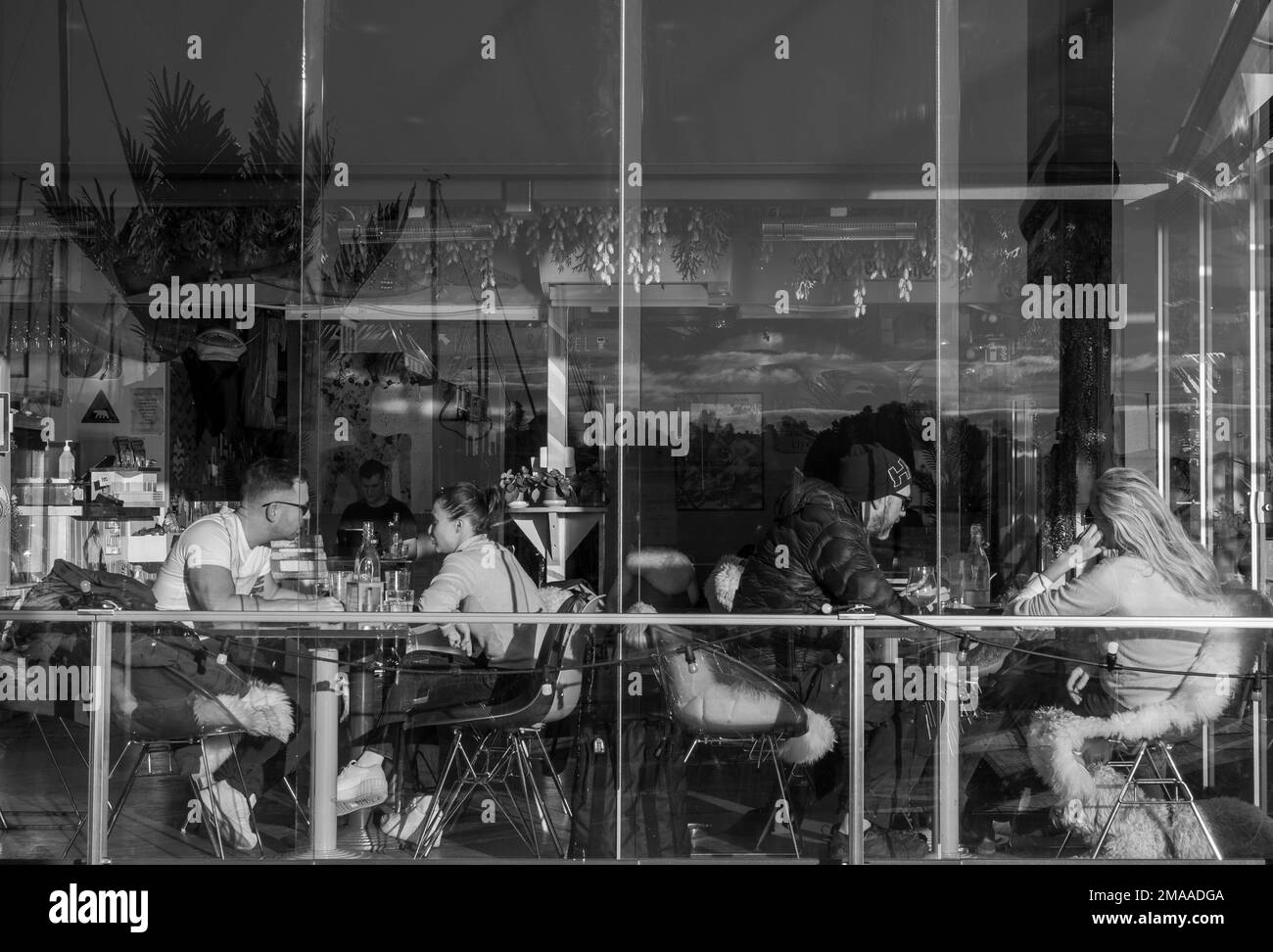Junge Paare sitzen an Tischen in der Kongen Marina Beach Bar & Restaurant, die an einem sonnigen Tag durch eine Glasfassade mit Reflexionen gesehen werden. Oslo, Norwegen. Stockfoto