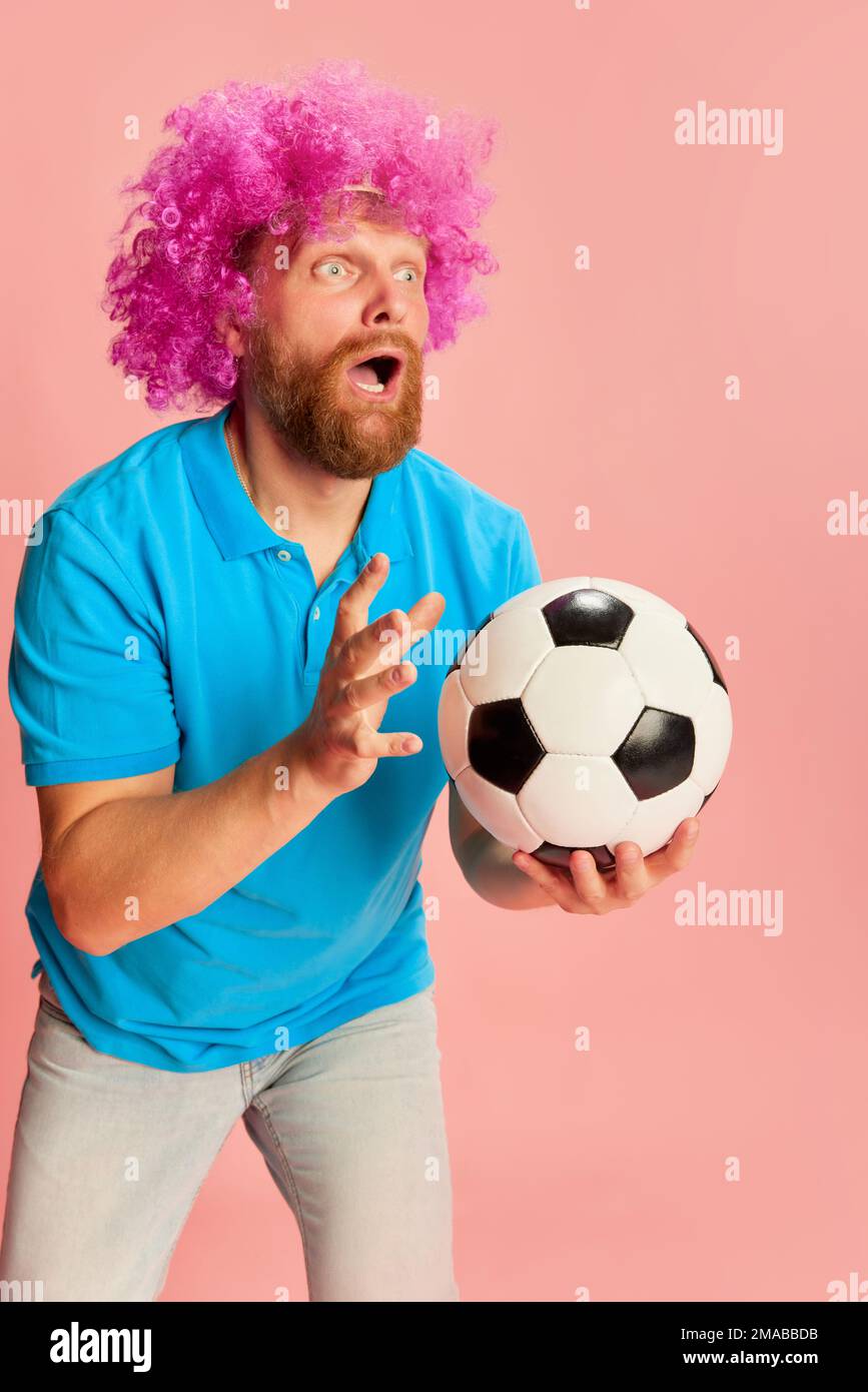 Emotionaler Fußballfan, junger weißer Mann mit lustiger Perücke, der sich ein Fußballspiel ansieht und Emotionen vor pinkfarbenem Hintergrund ausdrückt. Sport, Fans, Fußball Stockfoto