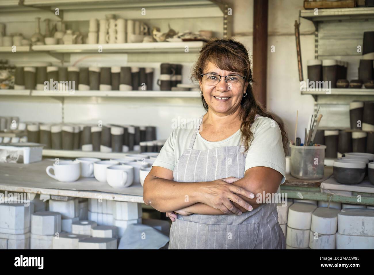 Erwachsene Frau Töpferin mit Brille und Arbeitsschürze lächelt und schaut in ihrem Studio in die Kamera. Stockfoto