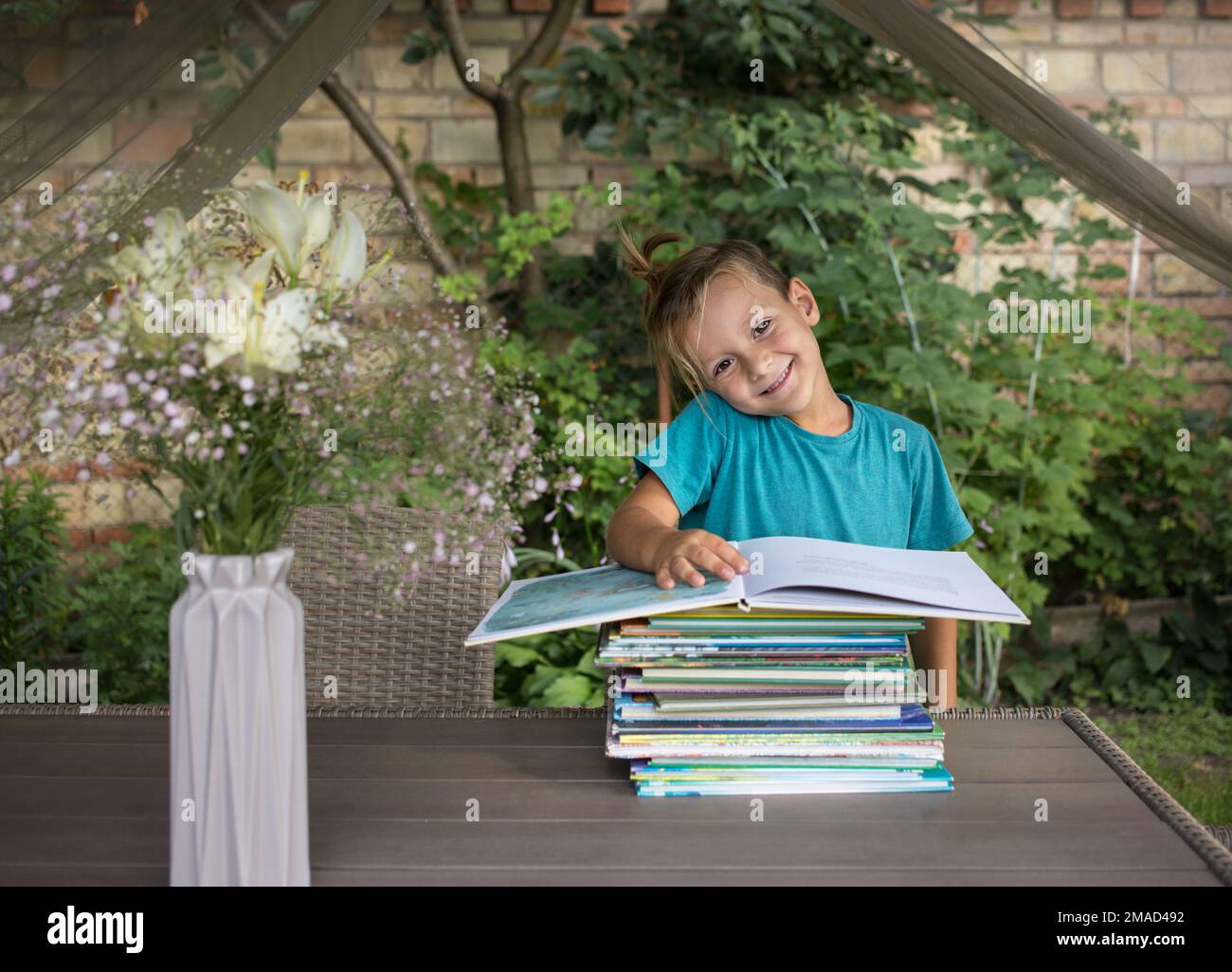 Lächelnder Vorschuljunge sitzt am Tisch, vor ihm ist ein Stapel Bücher. Bücher lesen, Kinderfantasien entwickeln, interessante Kindheit. Entzückend Stockfoto