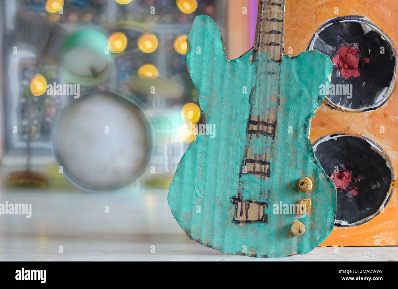 Schmieriges Pappmodell mit Gitarre und Lautsprecherbox, Synthesizer und Trommelkit verschwommen im Hintergrund, Musik, Performance, Instrumentenkonzept Stockfoto