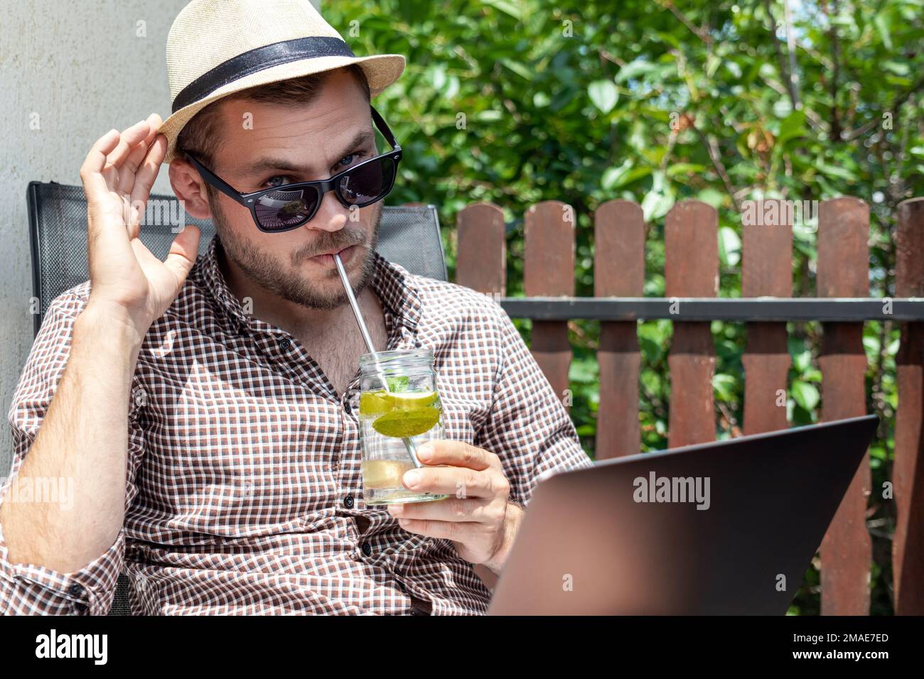 Ein gutaussehender Mann mit Sonnenbrille und Strohhut, der frisches Wasser mit Limette trinkt, während er im Sommer mit seinem Laptop auf der Terrasse sitzt. Stockfoto