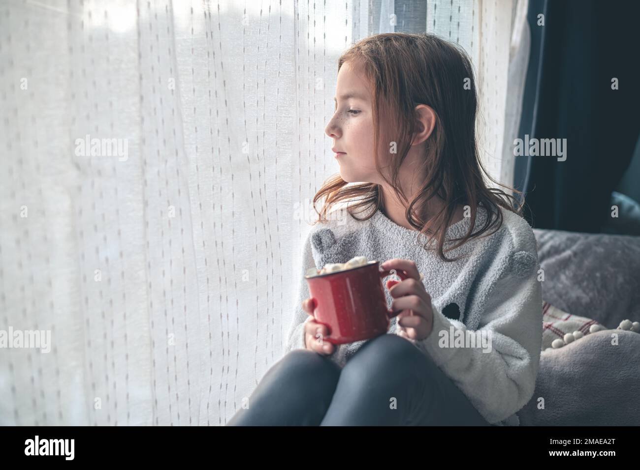 Ein süßes Mädchen auf dem Fensterbrett hält eine rote Tasse Kakao und Marshmallow und schaut aus dem Fenster. Stockfoto