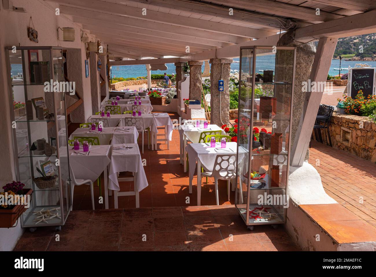 Restaurant-Mittagstische im Schatten eines hölzernen Vordachs mit buntem Blick auf die Straße und das Meer im Einkaufsviertel Bijou Sea Front in Baia Sardinia. Stockfoto